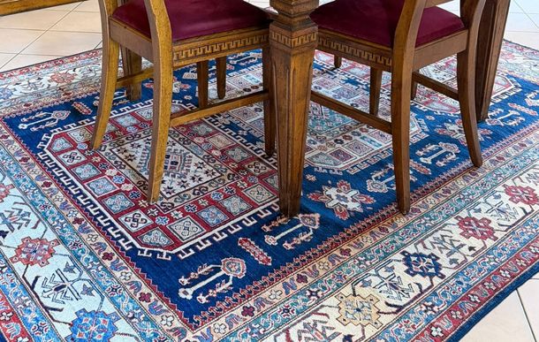 Dettaglio di un tavolo e sedie in legno con sedili in velluto bordeaux posizionati su un tappeto persiano tradizionale con motivi blu, rossi e crema.